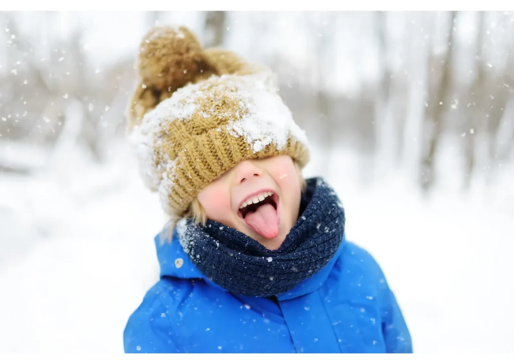 Child in a knitted hat and blue jacket sticking out their tongue in the snow