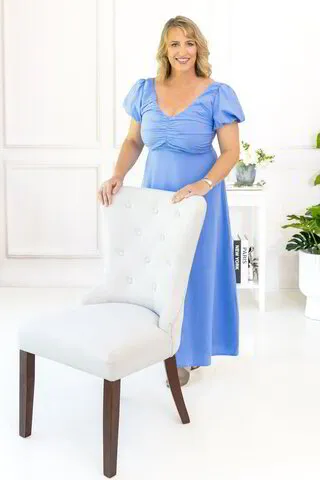 Emma Walker in a blue dress standing behind a cream armchair