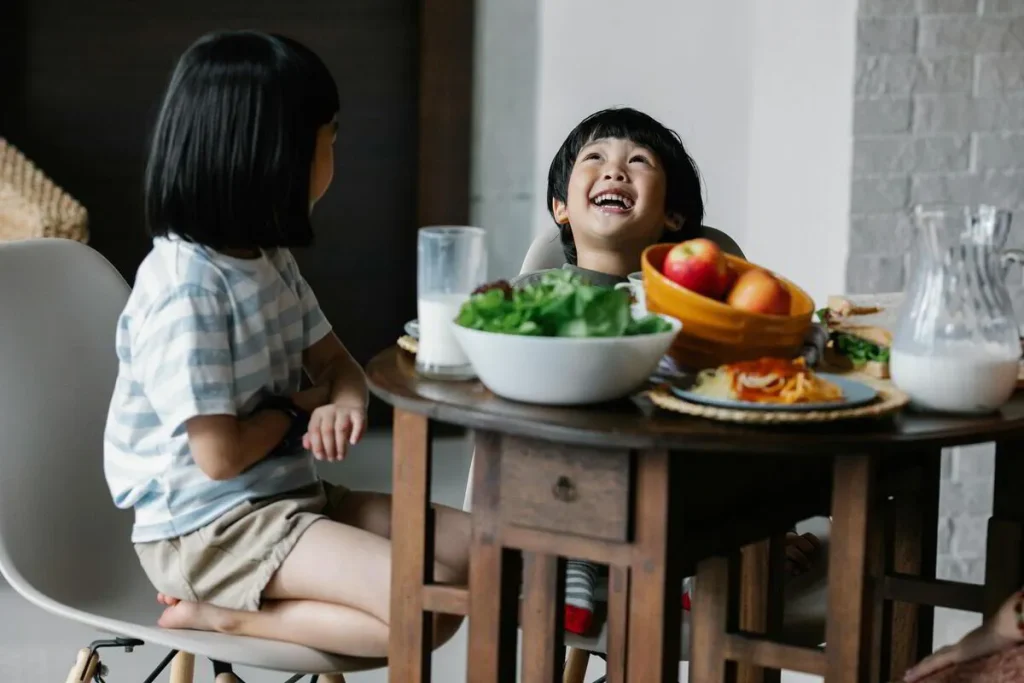 Girl at a dining table with a salad bowl and fruit while another child laughs beside her