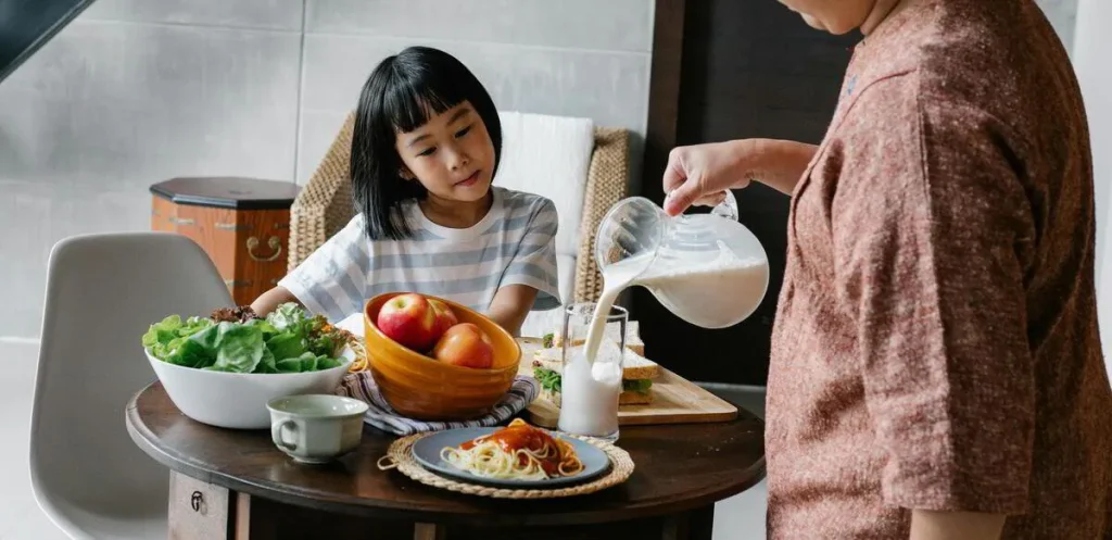 Girl at a table with a salad bowl and apples while an adult pours milk