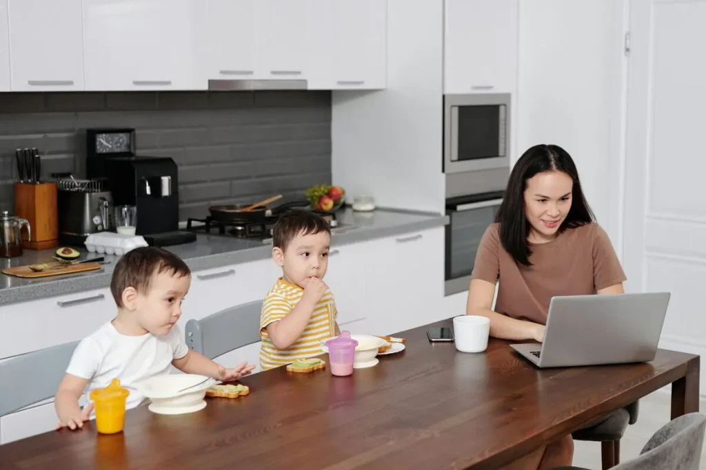Mother working on a laptop at the kitchen table with two toddlers beside her