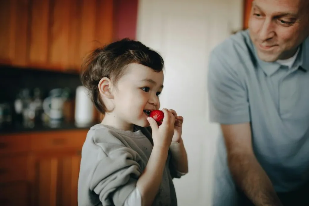 Toddler eating a cherry tomato in the kitchen beside an adult